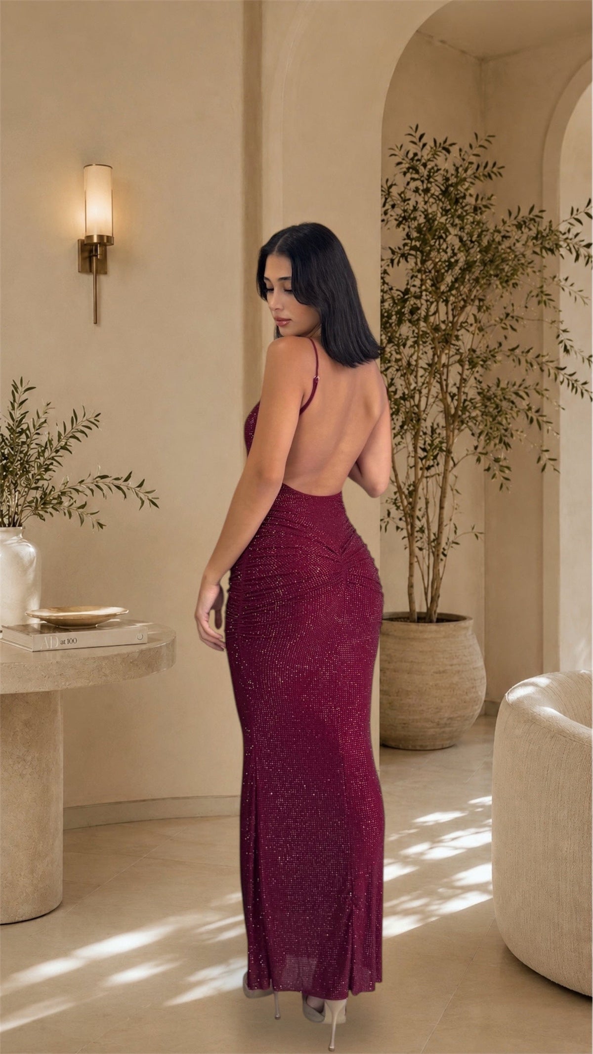 A woman wearing a sparkling burgundy open-back evening gown, photographed from behind in a warm, sunlit beige interior with plants and architectural arches.