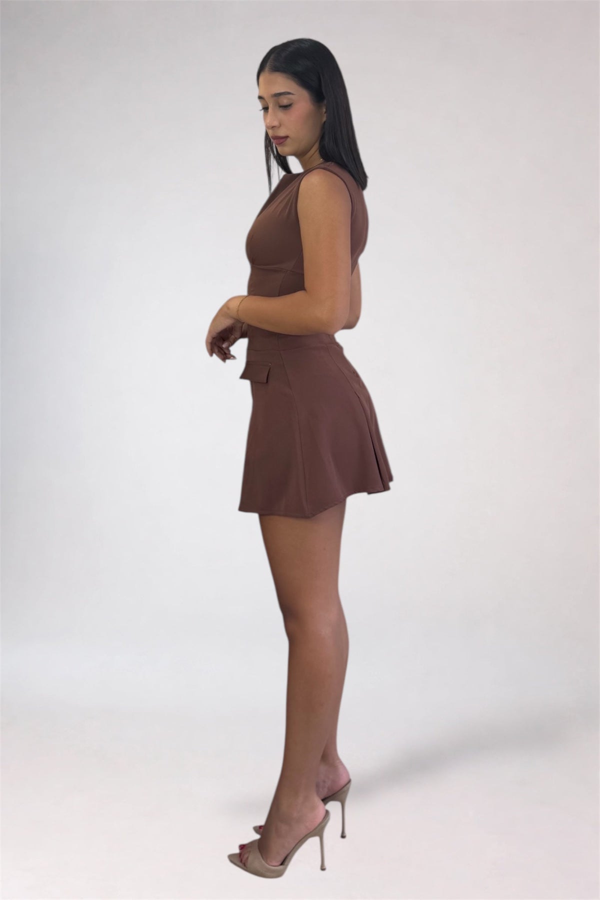 A woman modeling a sleeveless brown mini dress with a high neckline, side tie detail, and flared skirt, paired with nude high-heeled sandals, photographed against a plain light gray studio background.