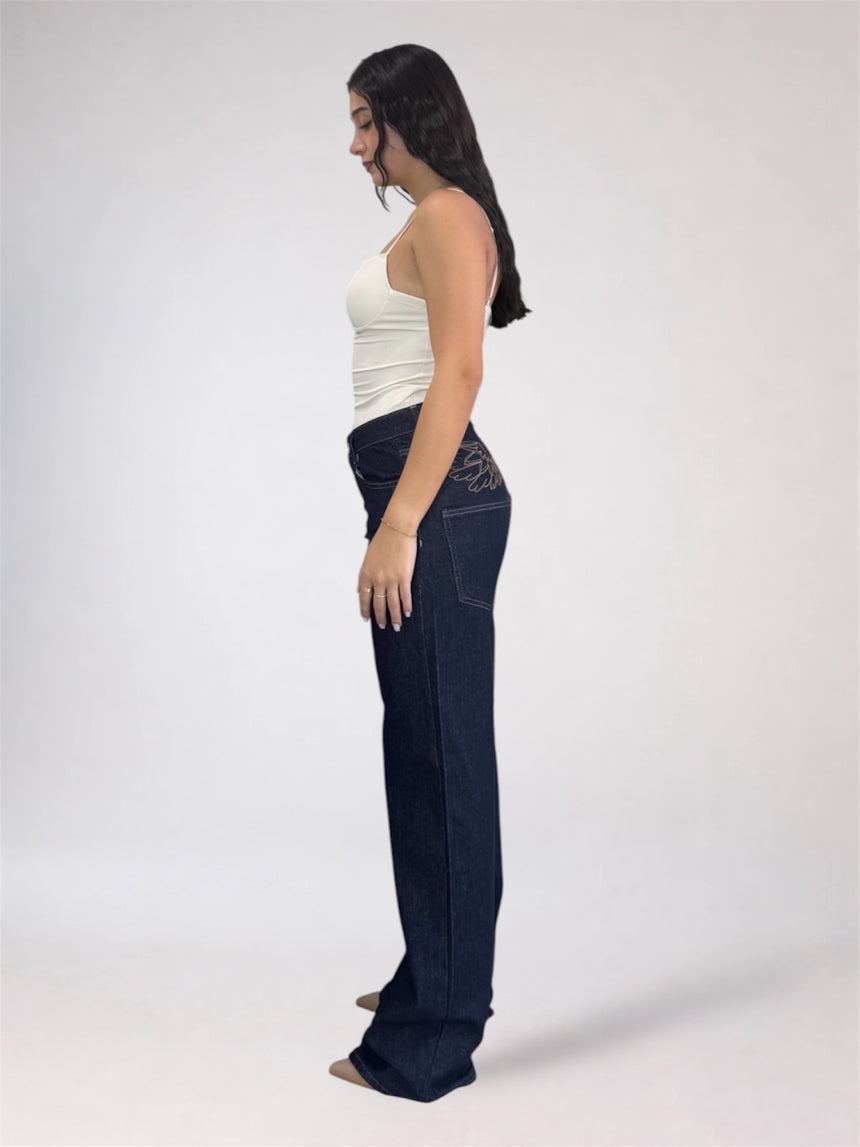 A woman shown in side profile wearing dark blue wide-leg jeans and a white tank top against a plain light gray studio background.