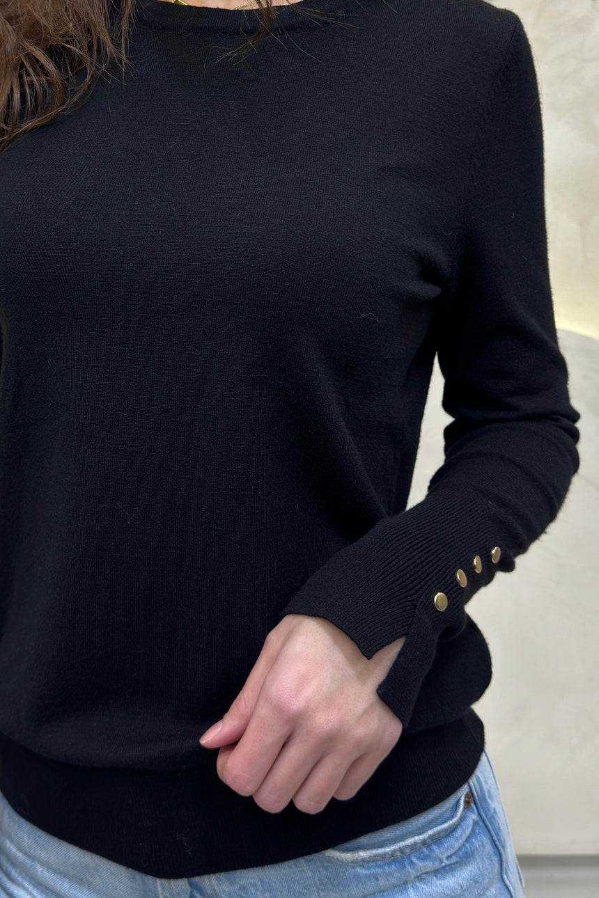 Black sweater with gold buttons on cuffs worn by a person, against a neutral background