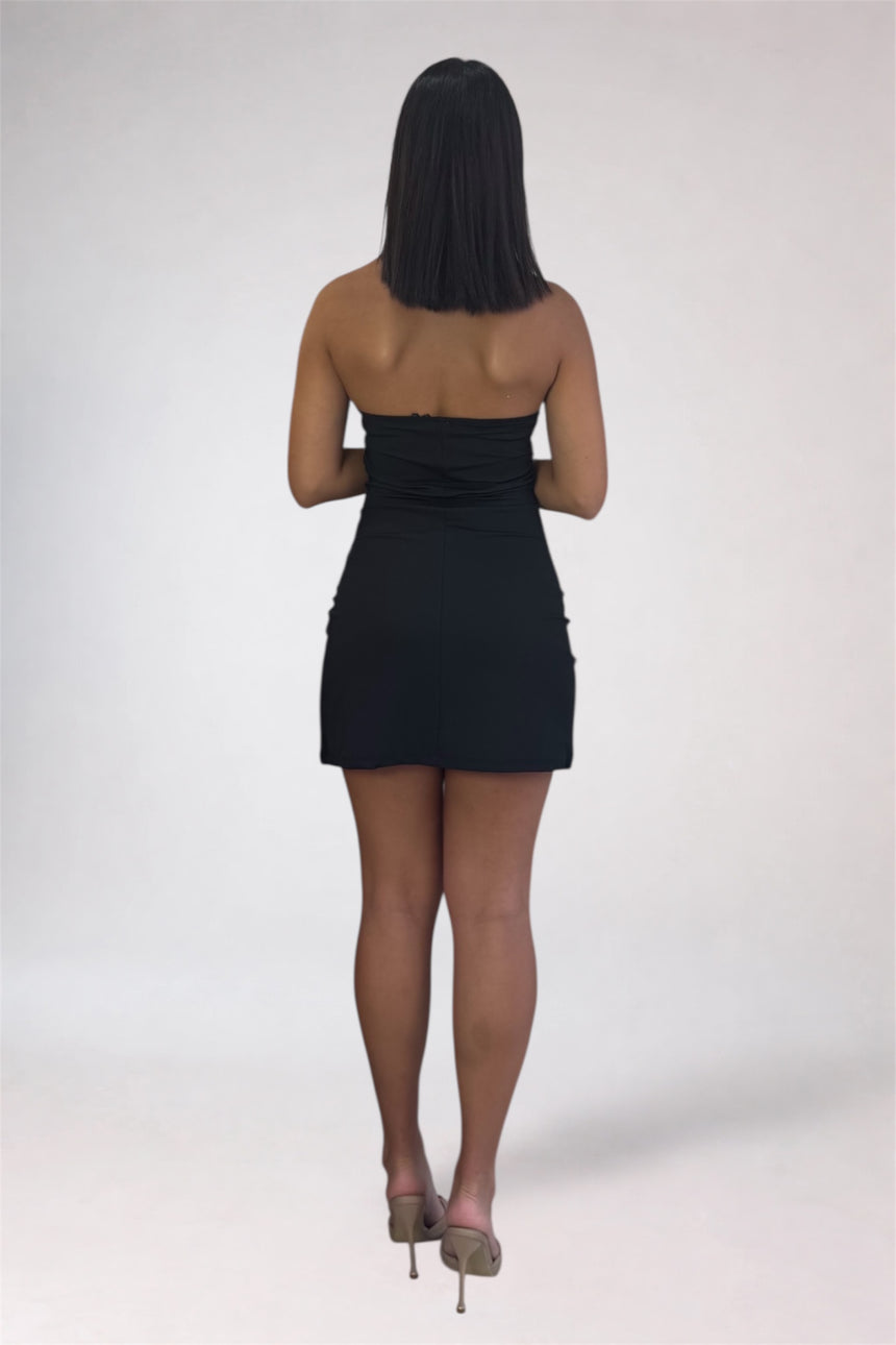 A woman viewed from behind wearing a strapless black mini dress and nude high heels against a plain light gray studio background.
