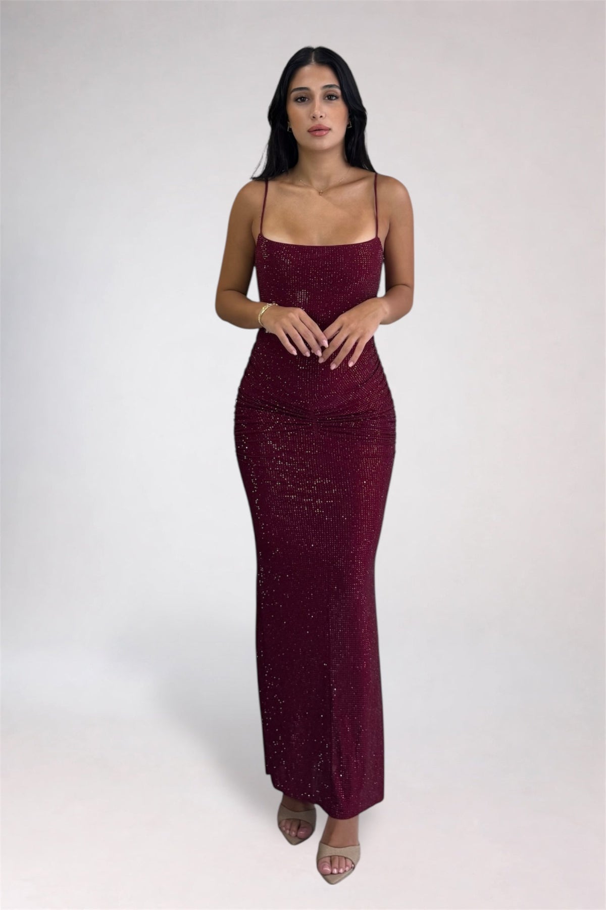 A woman modeling a floor-length, bodycon burgundy sequin maxi dress with spaghetti straps and a square neckline, paired with clear high-heeled sandals, against a light gray studio background.
