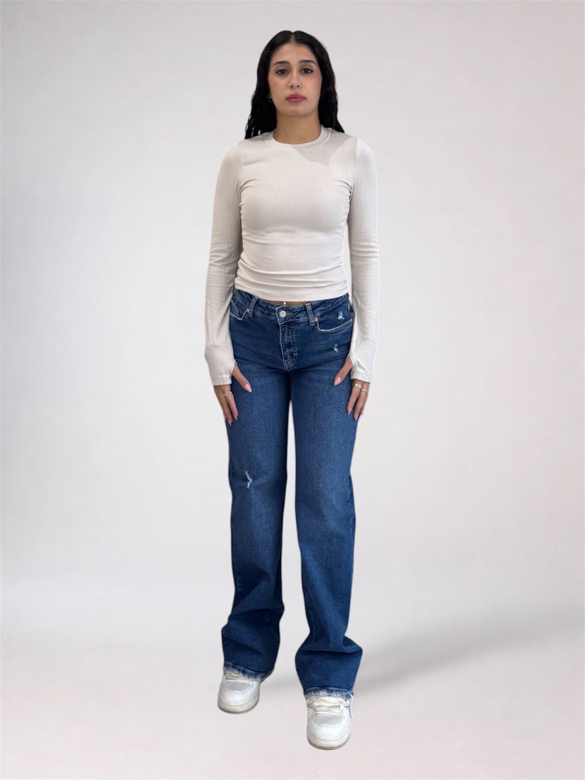 A woman modeling a fitted off-white long-sleeve top, blue straight-leg jeans, and white sneakers against a plain light gray studio background.