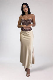 A woman modeling a strapless formal gown with a brown lace bustier-style bodice and a flowing champagne satin skirt with a side slit, photographed against a light gray studio background.