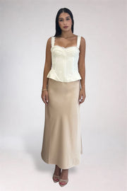 A woman modeling a two-piece outfit consisting of a cream corset-style camisole top and a matching beige maxi skirt, photographed against a plain light gray studio background.