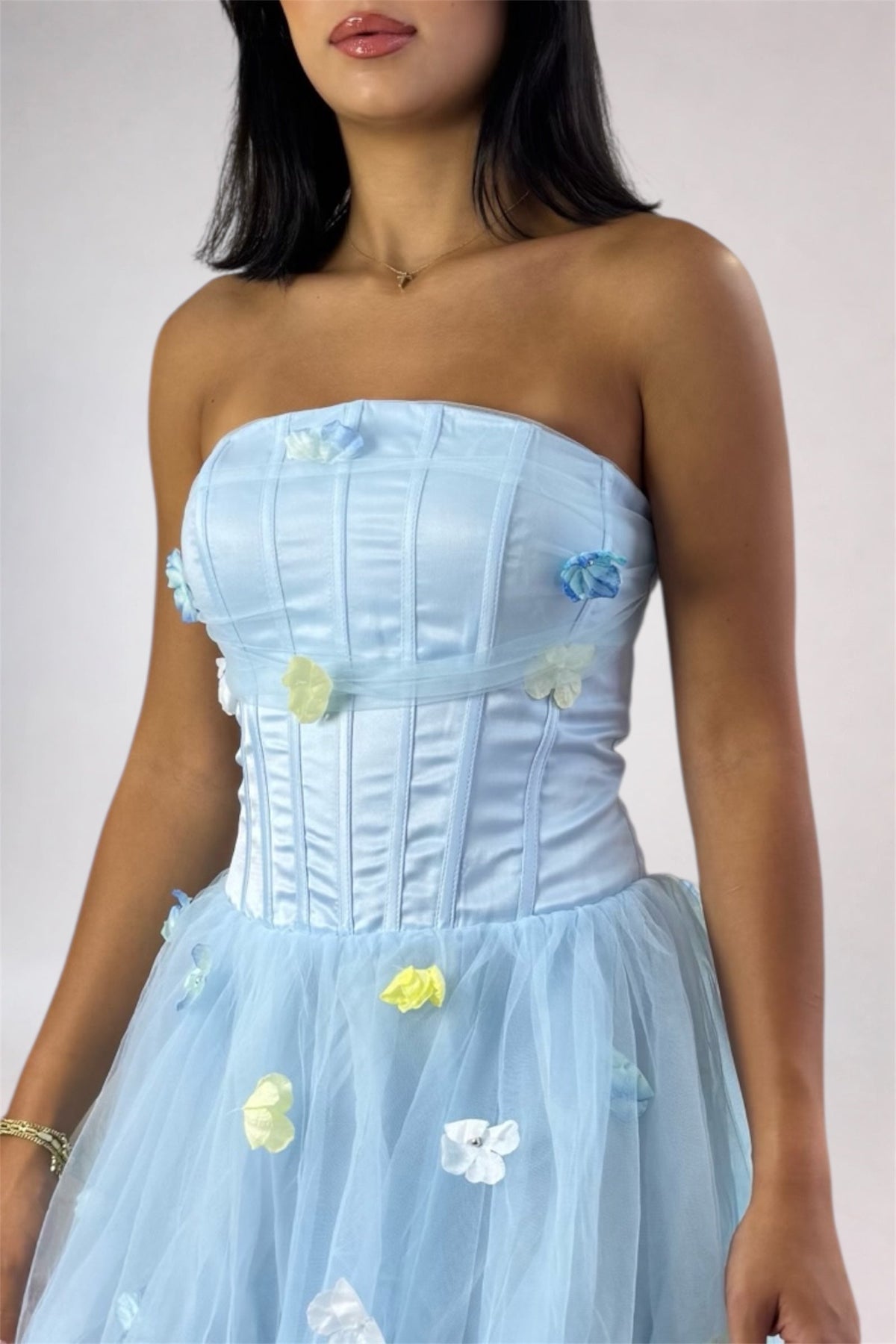A light blue strapless corset dress with a flared tulle skirt, decorated with small 3D fabric flowers in pastel blue, yellow, and white. The dress is shown on a model against a plain light gray studio background.