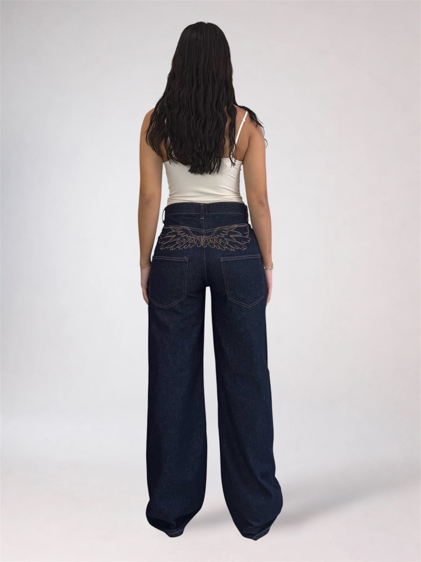 A woman viewed from behind wearing dark blue wide-leg jeans with a silver embroidered wing design on the back waistband, paired with a white spaghetti-strap tank top, against a light gray studio background.