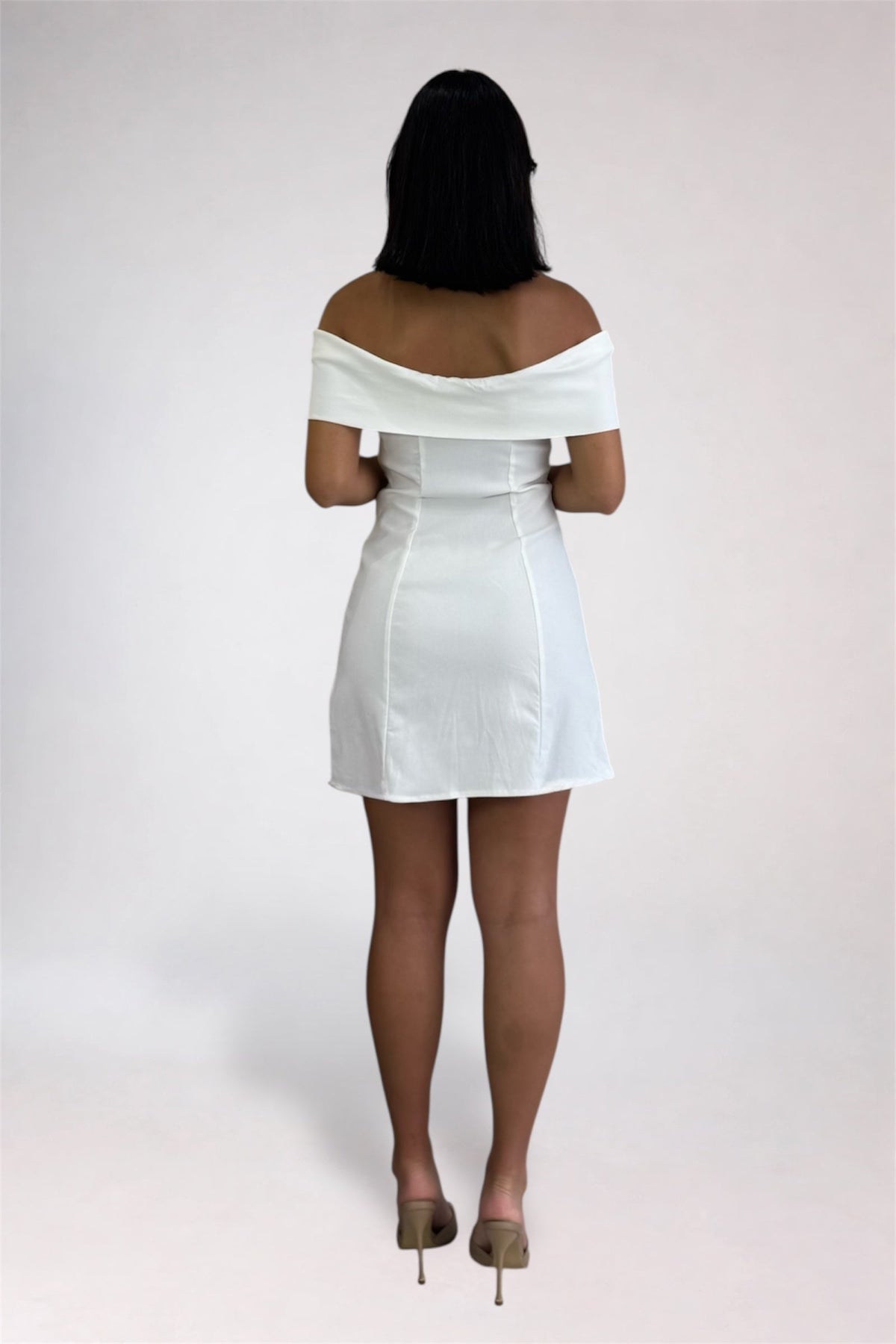 A woman viewed from behind wearing an off-the-shoulder white mini dress with structured shoulders and taupe high heels, standing against a light gray studio background.