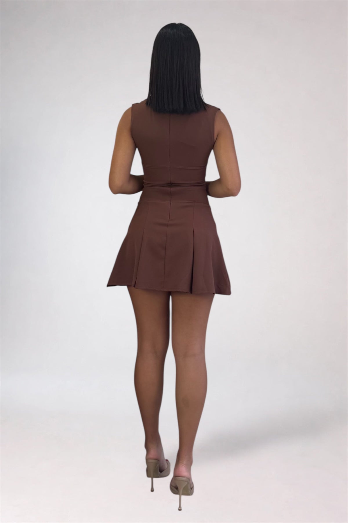 A woman viewed from behind wearing a sleeveless brown mini dress with a flared skirt and matching belt, paired with nude tights and beige heels, against a light gray studio background.