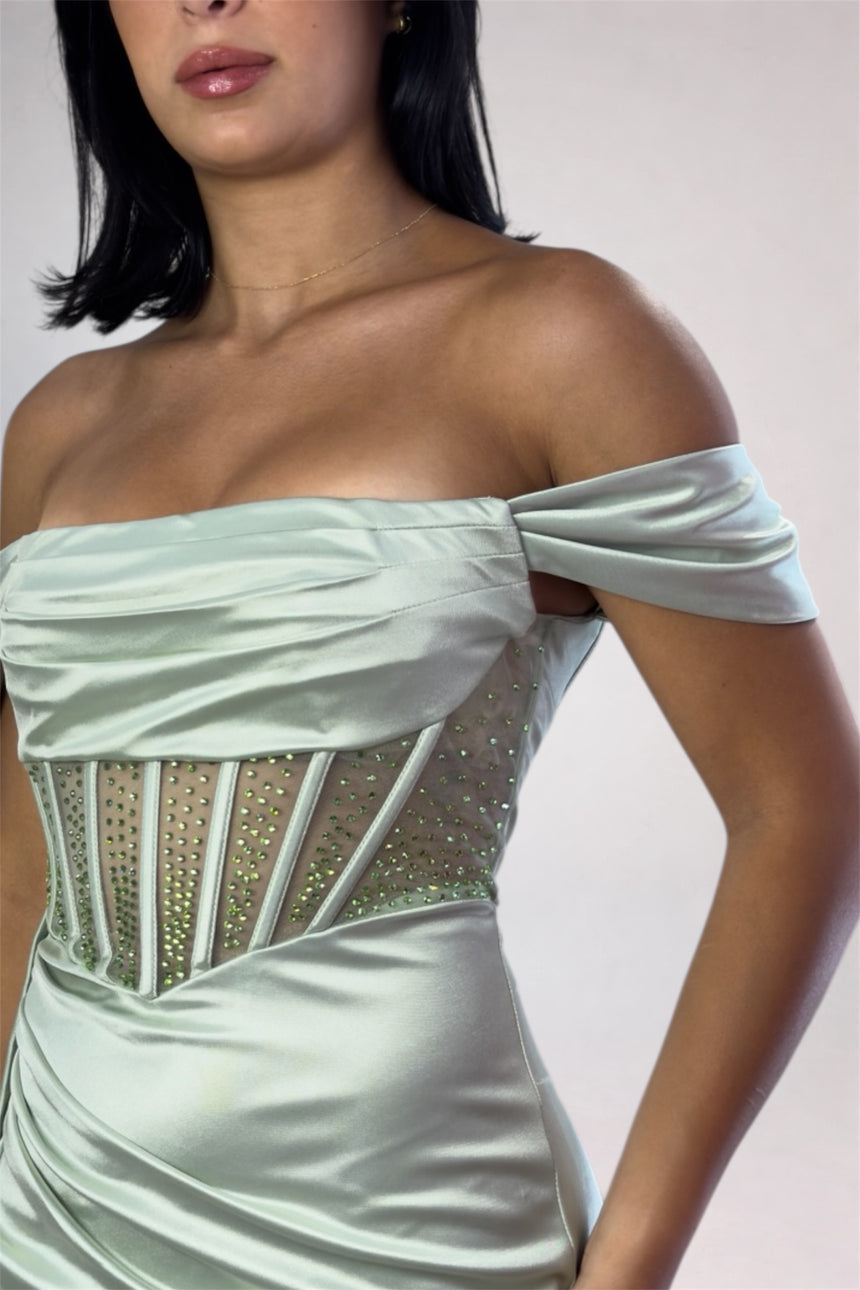 An off-the-shoulder mint green satin dress with a ruched bodice, sheer mesh panels, and gold rhinestone trim, shown on a model against a light gray background.