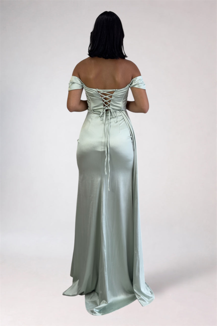 A woman viewed from the back wearing a floor-length off-the-shoulder satin gown with lace-up back detailing, photographed against a plain light studio background.