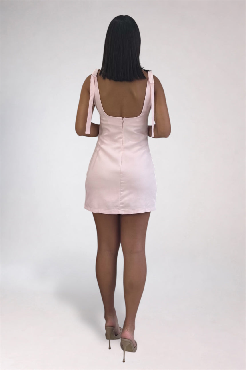 A woman viewed from behind wearing a light pink mini dress with shoulder straps and a low open back, paired with nude high heels, against a plain light gray studio background.