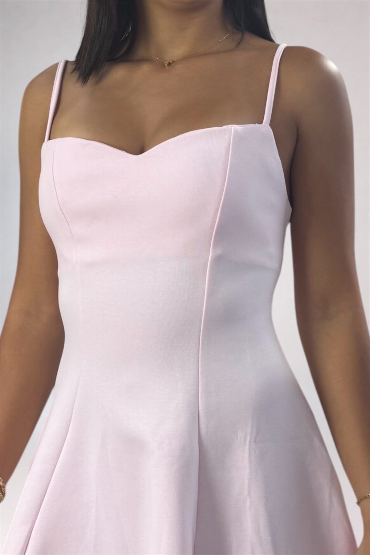 A woman wearing a light pink spaghetti-strap dress with a sweetheart neckline and a flared skirt, shown from the shoulders to the hips against a plain light gray background.