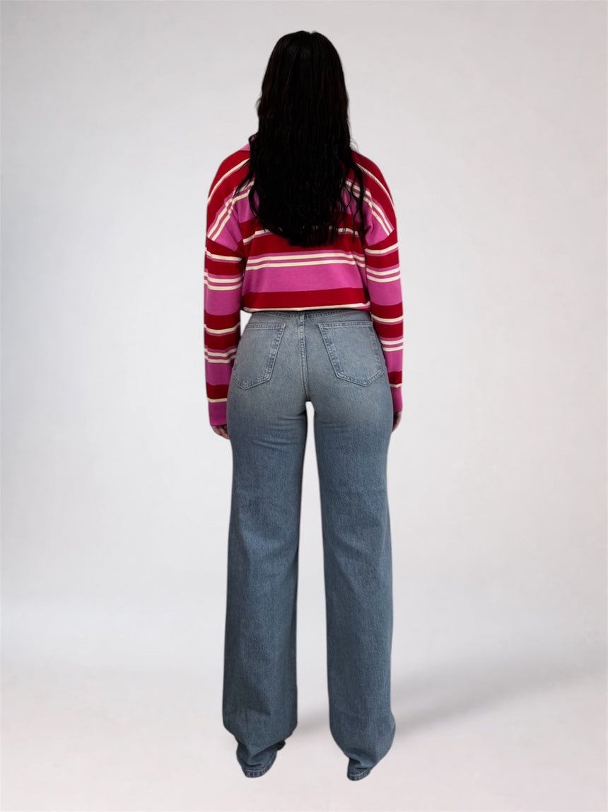 A woman viewed from behind wearing a red, pink, and white striped long-sleeve crop top and light blue wide-leg jeans, standing against a light gray studio background.