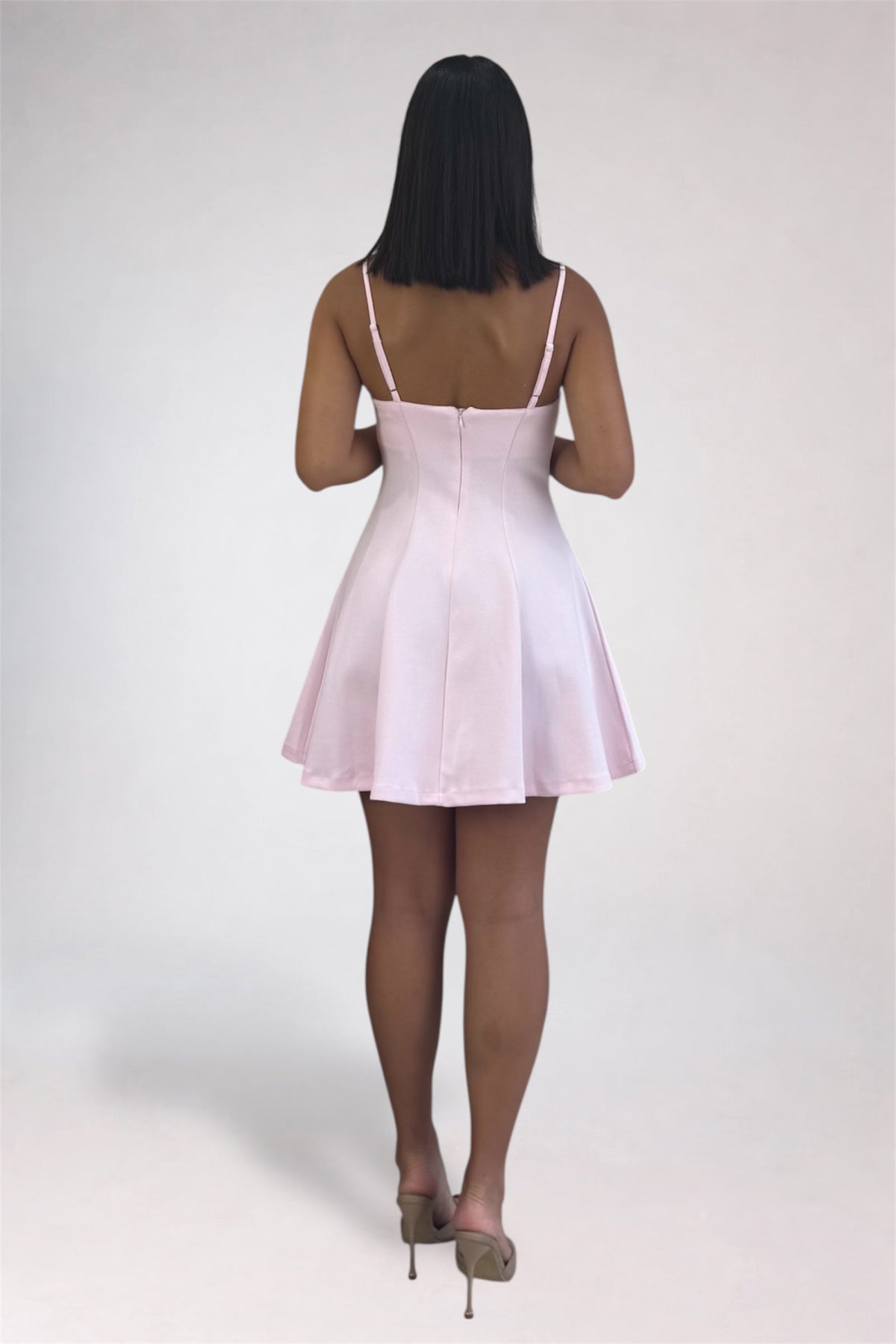 A woman viewed from behind wearing a light pink spaghetti-strap mini dress with a fitted bodice and flared skirt, paired with nude high heels, against a plain light gray studio background.