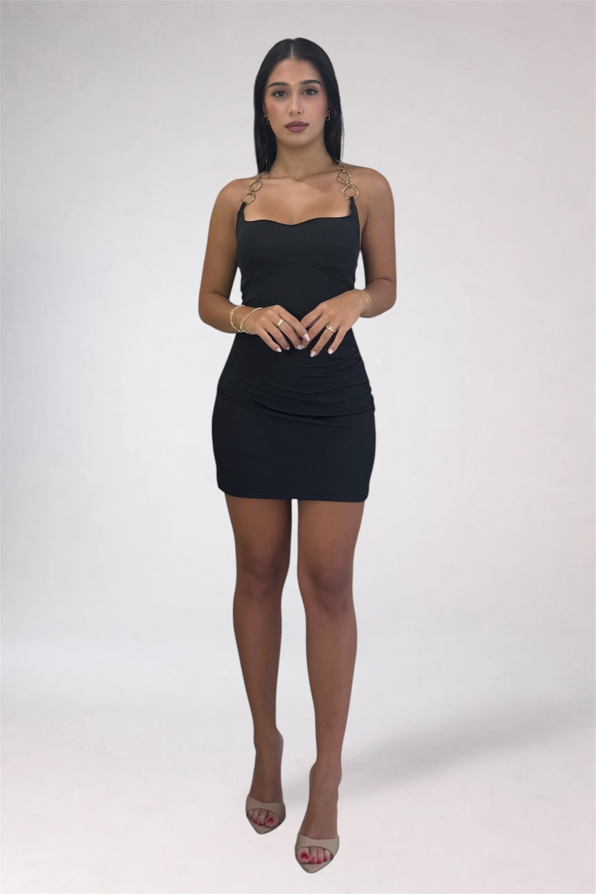 A woman modeling a black bodycon mini dress with shoulder cutouts and gold chain details on the straps, paired with nude high-heeled sandals, against a light gray studio background.
