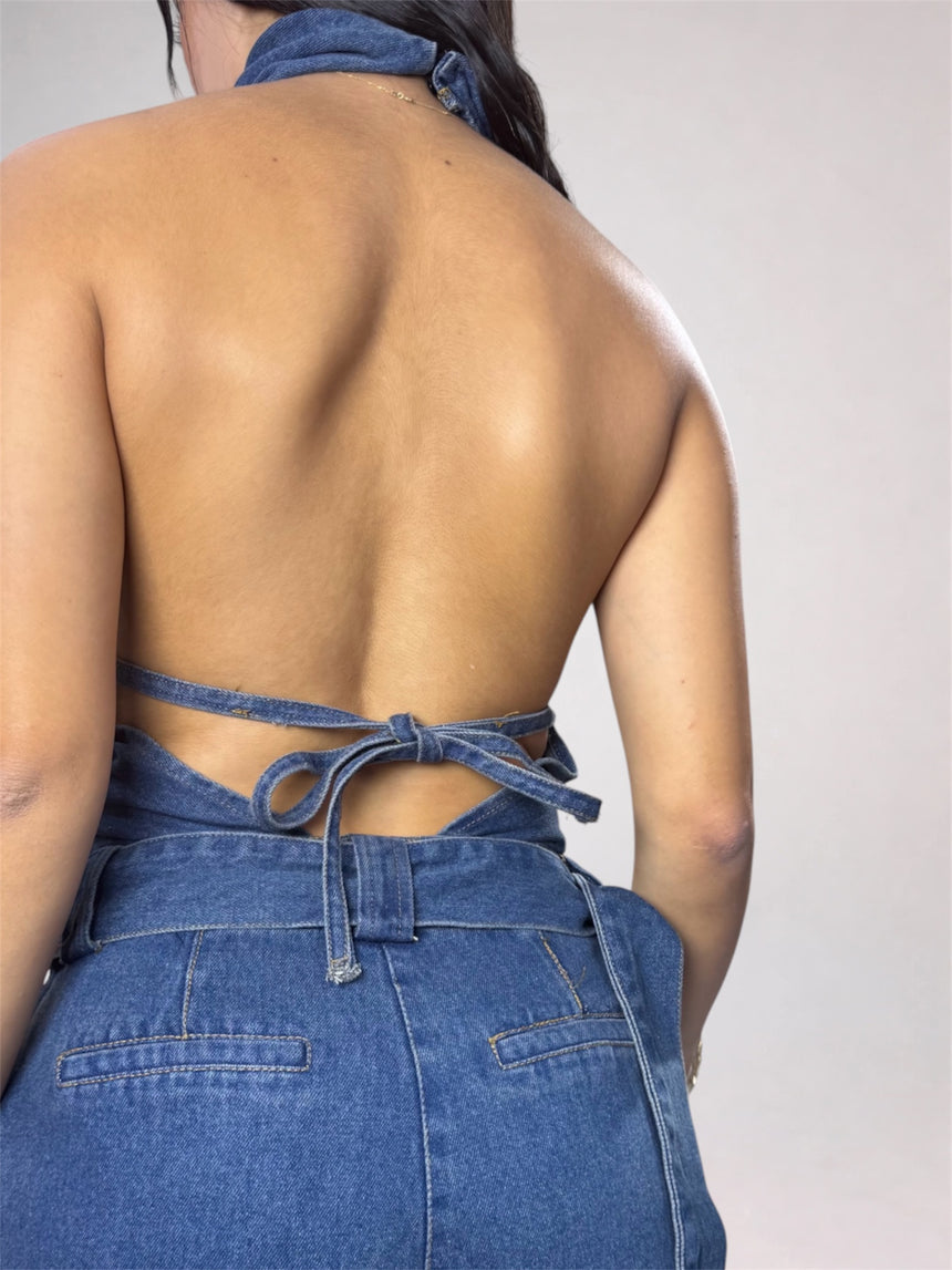 Rear view of a woman wearing a blue denim halter top and matching denim bottoms against a plain light gray studio background.