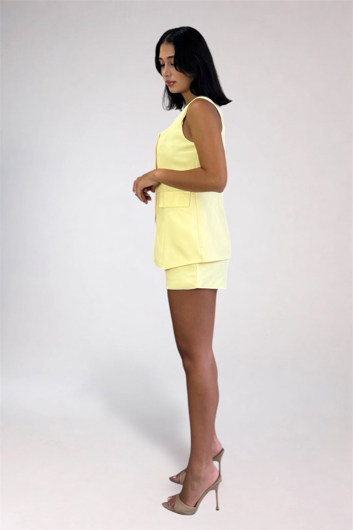 A woman modeling a pale yellow sleeveless vest and matching shorts set, standing in profile against a light gray studio background.