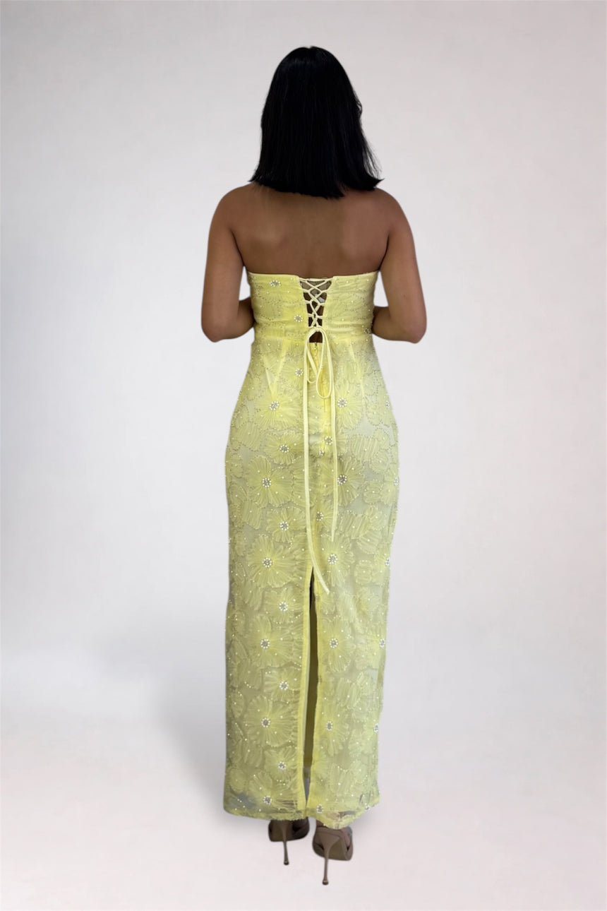 A woman viewed from the back wearing a strapless pale yellow lace dress with a lace-up back and front slit, standing against a plain light gray studio background.
