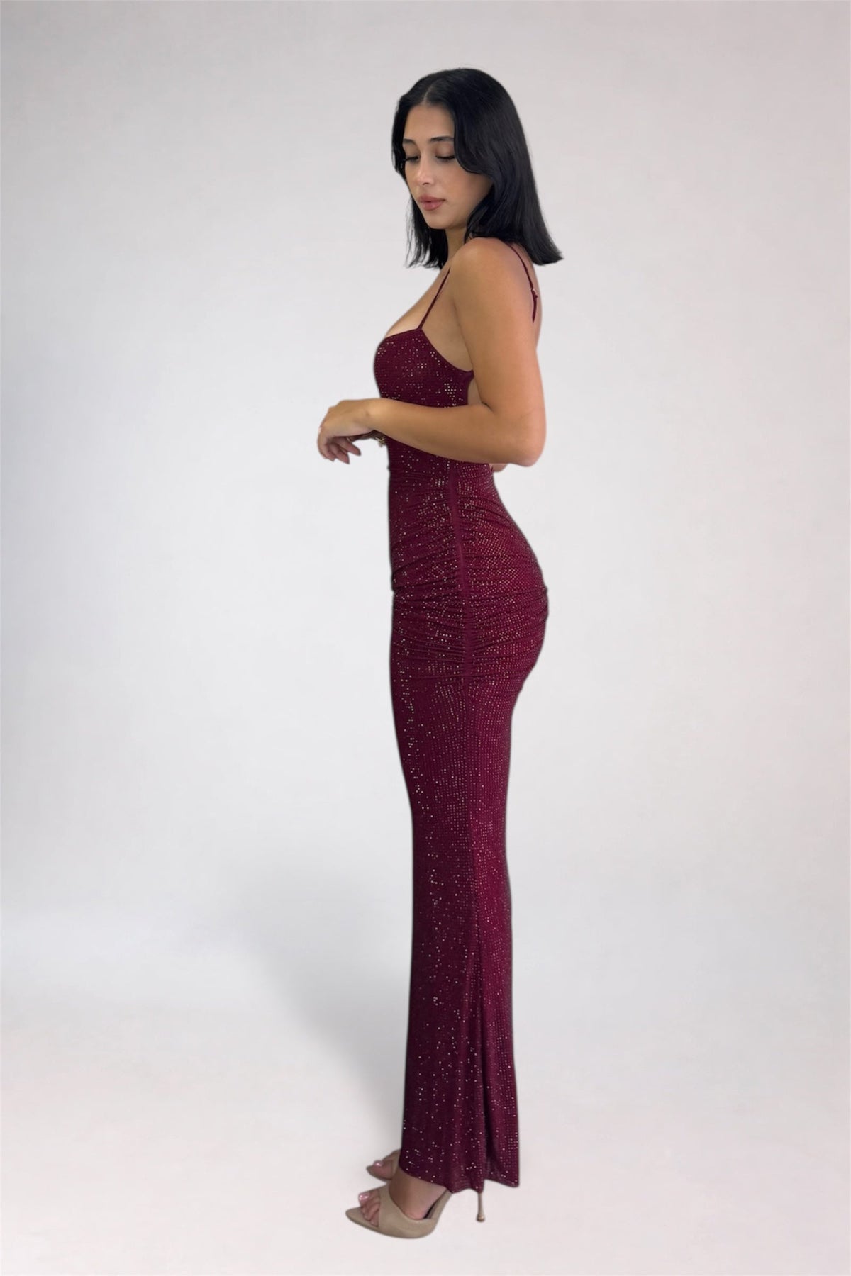 A woman modeling a sleeveless, bodycon maxi dress covered in sparkling sequins. The dress is a deep burgundy/wine color, shown in profile against a plain light gray studio background.