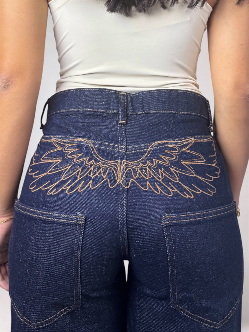 Rear view of a person wearing dark blue denim jeans with a large embroidered wing design across the back pocket, paired with a light beige top against a plain light background.