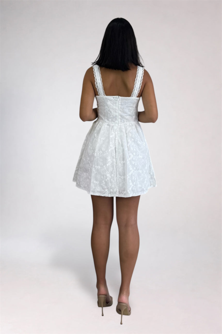 A woman viewed from the back wearing a short white lace dress with wide straps and a flared skirt, standing against a plain light gray studio background.