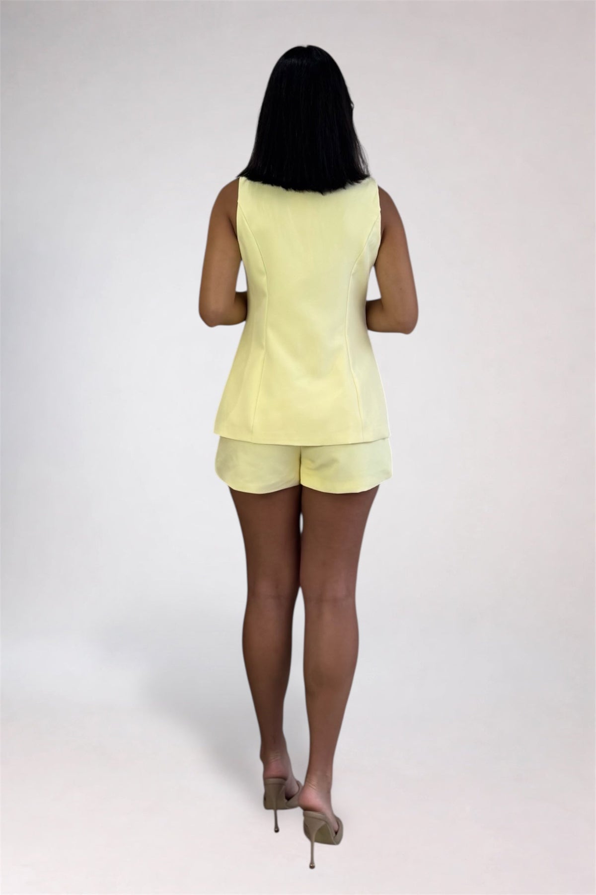 A woman viewed from behind wearing a pale yellow sleeveless top and matching shorts, standing against a plain light gray studio background.
