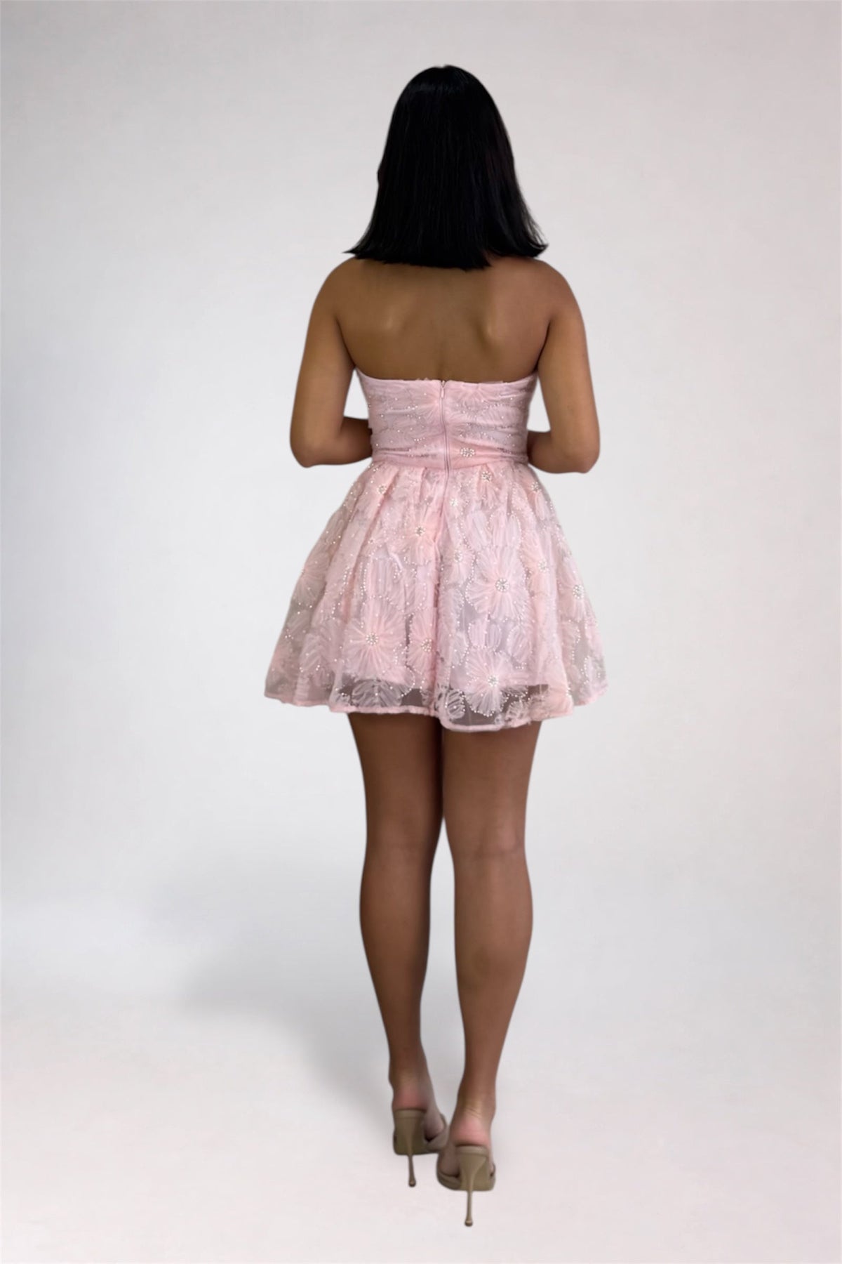 A woman viewed from behind wearing a light pink strapless lace mini dress with a flared skirt and nude high heels, standing against a plain light gray studio background.