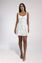 A woman modeling a white sleeveless mini dress with a small red and green floral print, standing against a light gray studio background.