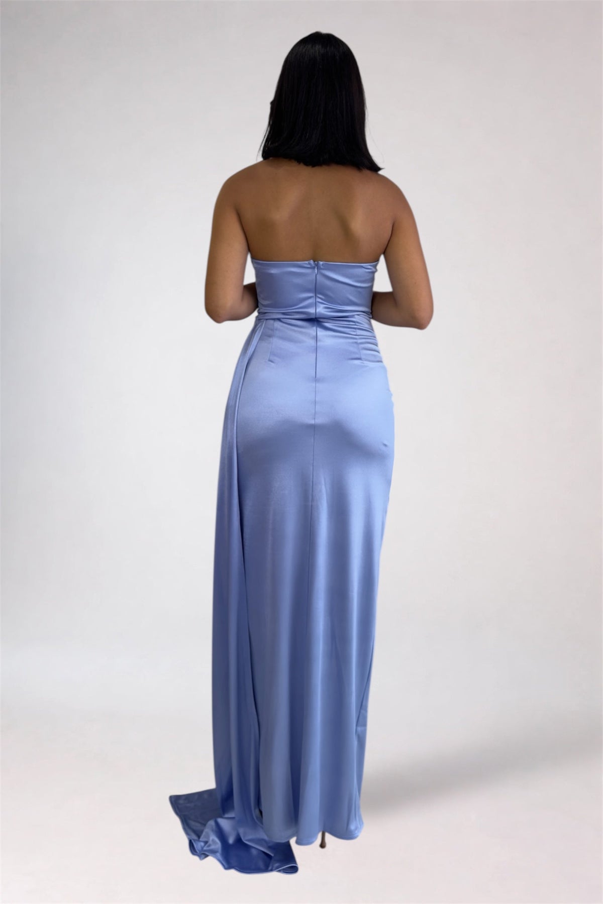 A woman viewed from the back wearing a strapless light blue satin maxi dress with a fitted waist and a long flowing skirt, photographed against a plain light gray studio background.