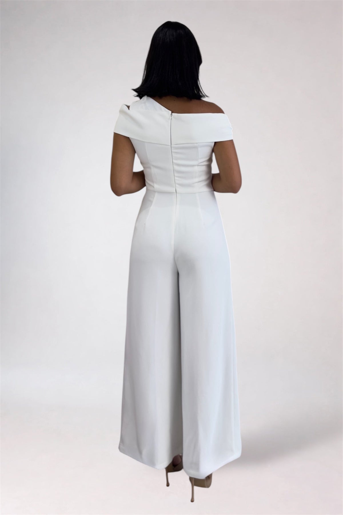 A woman viewed from the back wearing a white off-the-shoulder wide-leg jumpsuit, standing against a plain light gray studio background.