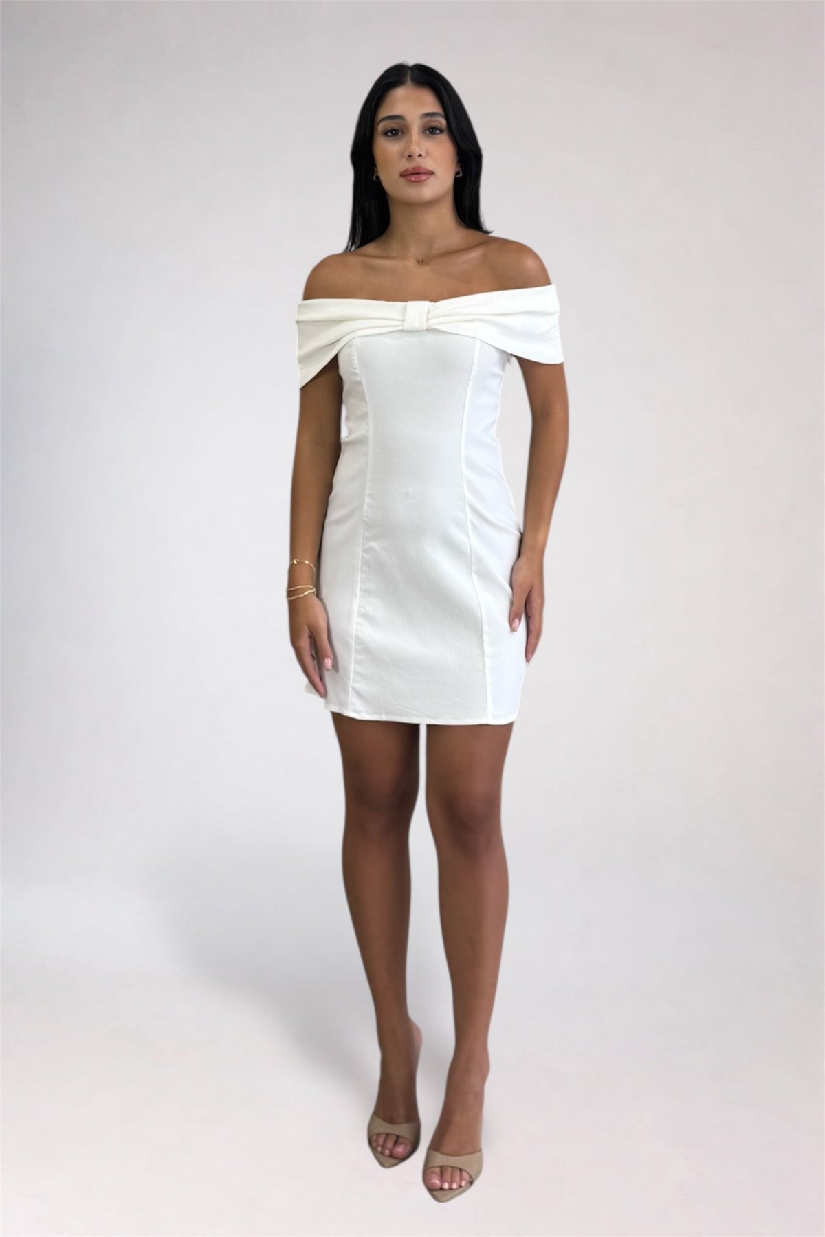A woman modeling an off-the-shoulder white mini dress with structured side seams and a bow detail at the neckline, photographed against a plain light gray studio background.