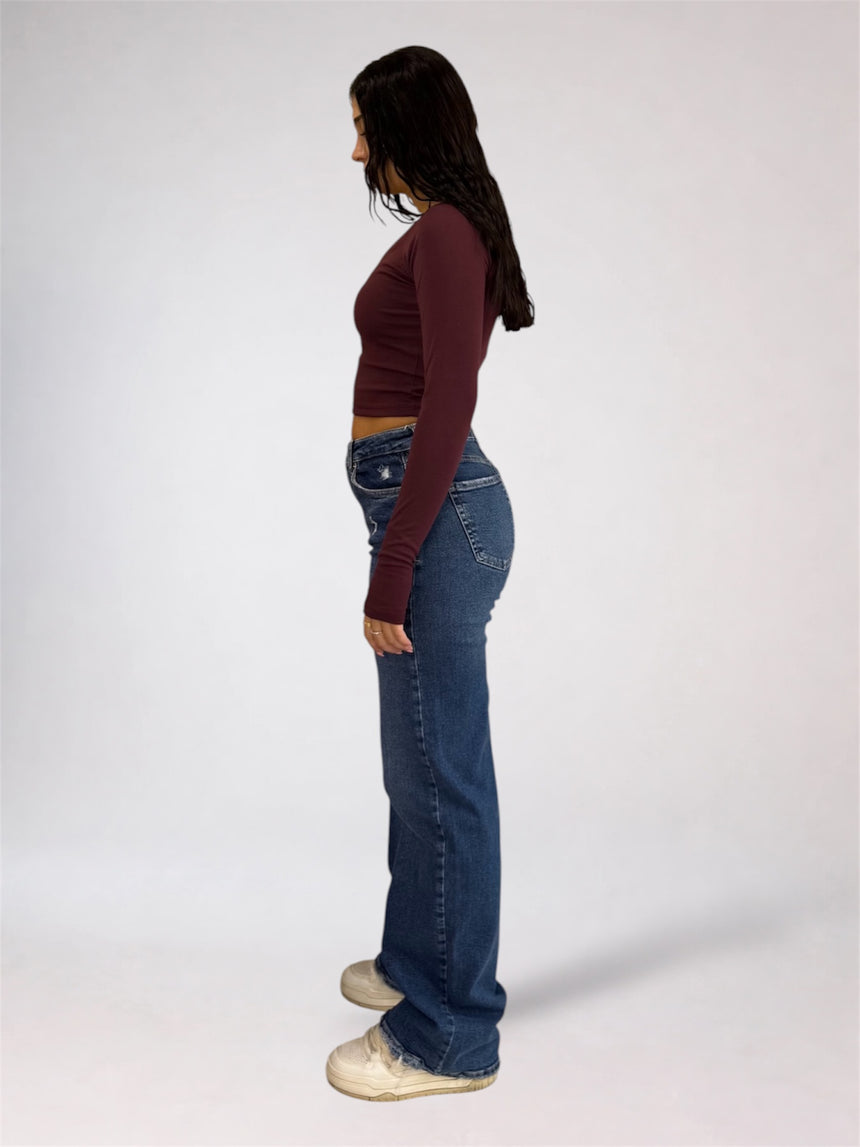 A woman shown in profile wearing a dark burgundy long-sleeve crop top, blue flare jeans, and off-white sneakers against a plain light gray studio background.