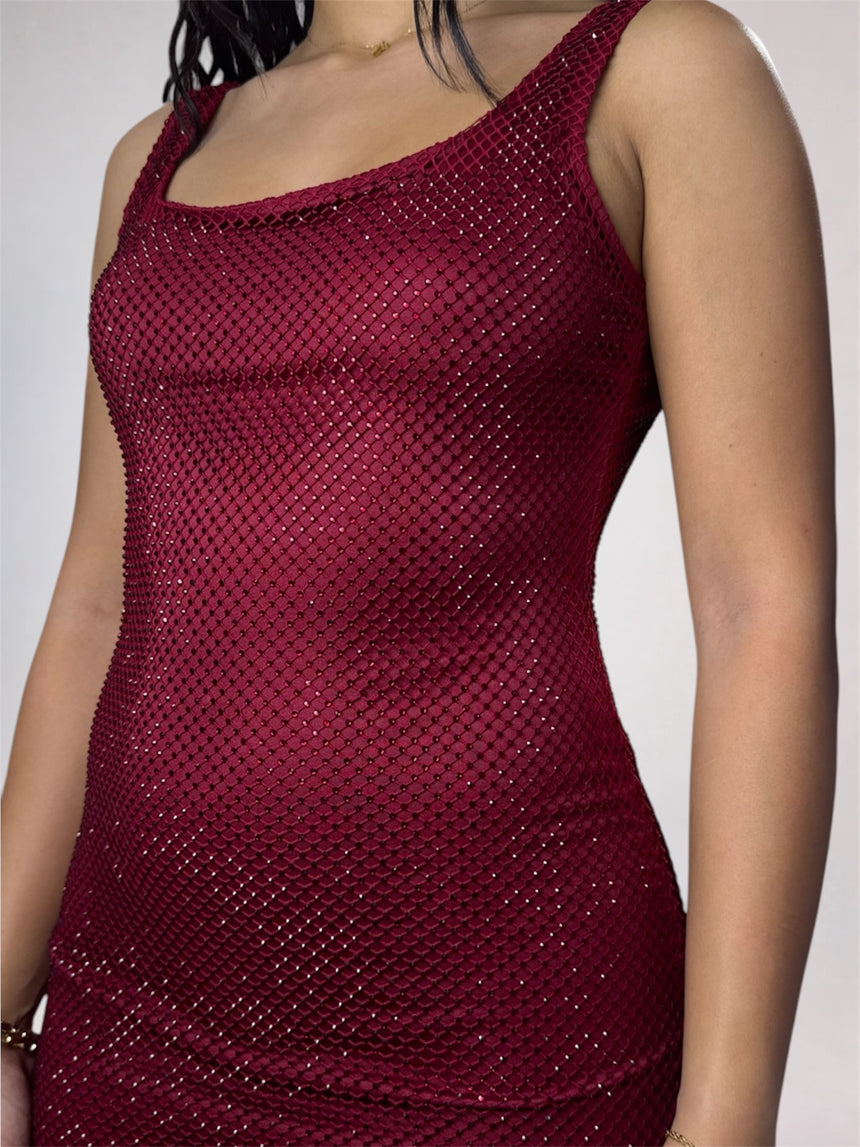 A close-up of a woman wearing a sleeveless scoop-neck dress covered in shimmering dark red sequins or mesh-like embellishment against a plain light gray background.