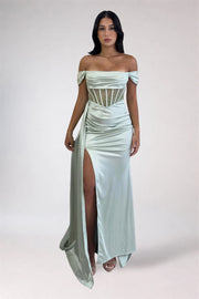 A woman modeling an off-the-shoulder sage green satin maxi gown with a high thigh slit and a corset-style cutout bodice, set against a plain light gray studio background.