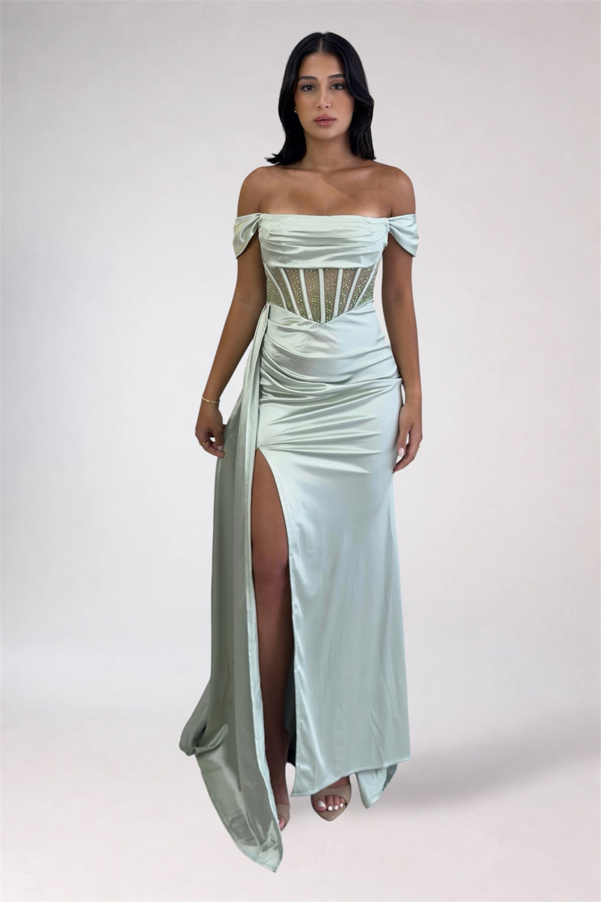 A woman modeling an off-the-shoulder sage green satin maxi gown with a high thigh slit and a corset-style cutout bodice, set against a plain light gray studio background.