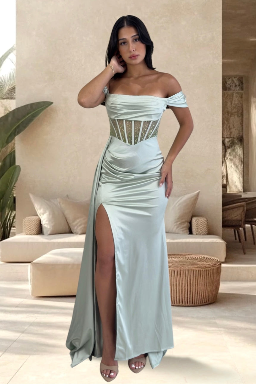 A woman models an off-the-shoulder light sage green satin gown with a corset-style bodice, ruched skirt, and high thigh slit in a warm, modern indoor setting.