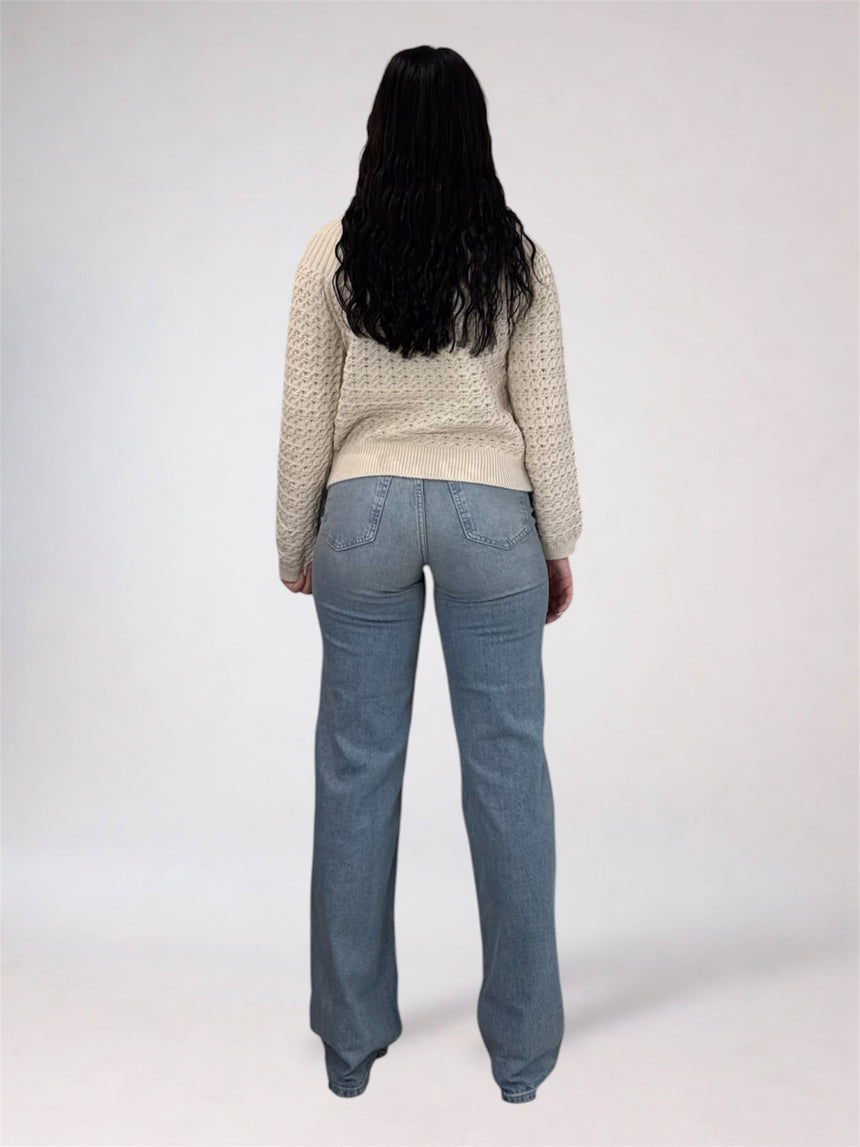 A woman viewed from behind wearing a cream cable-knit sweater and light blue straight-leg jeans against a plain light gray studio background.