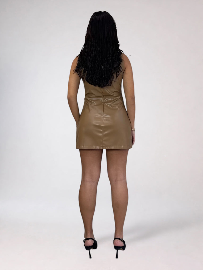 A woman viewed from behind wearing a sleeveless, mini-length brown faux-leather dress and black high-heeled sandals against a plain light gray studio background.