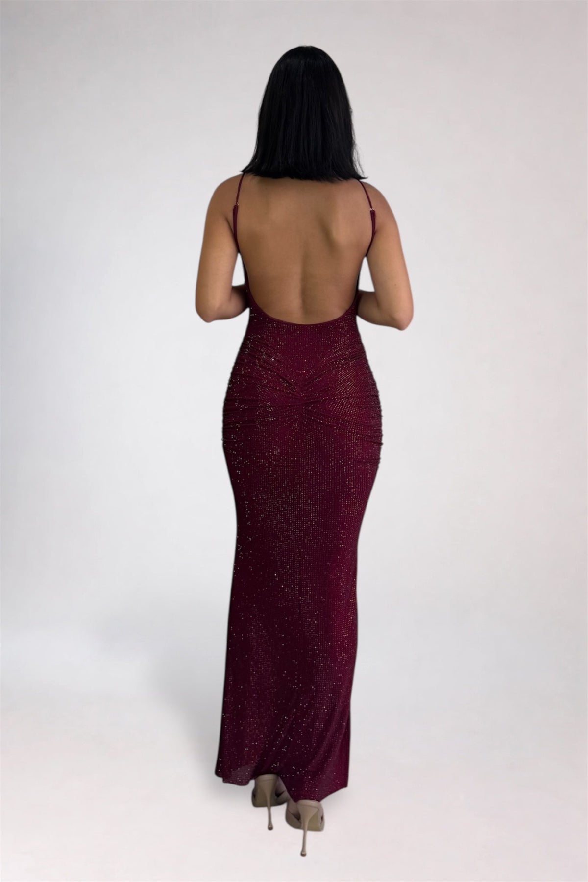 A woman viewed from the back wearing a floor-length, open-back burgundy sequined gown with thin straps and nude heels, standing against a plain light gray studio background.