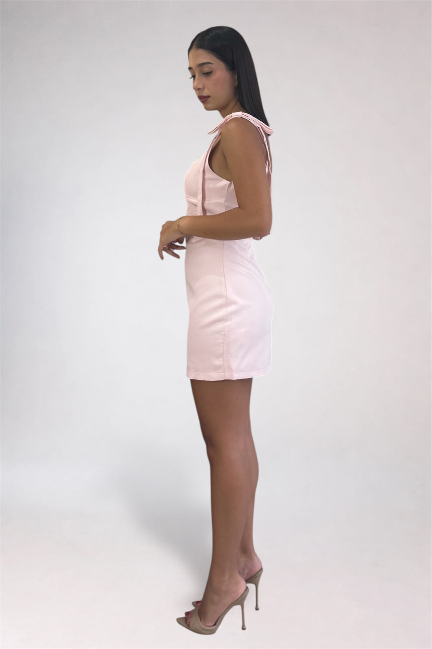A woman modeling a light pink sleeveless mini dress with tie-shoulder straps, paired with clear high-heeled sandals, standing against a plain light gray studio background.