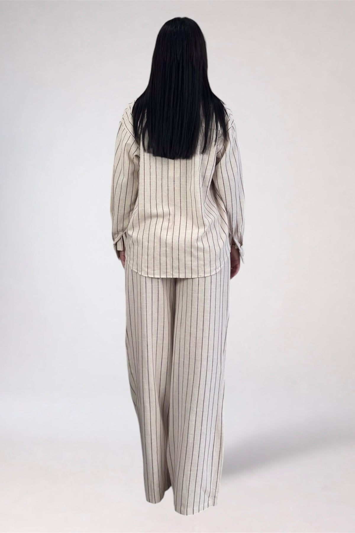 A woman viewed from behind wearing a matching two-piece pajama set with vertical stripes, consisting of a long-sleeve top and wide-leg pants, against a plain light gray background.