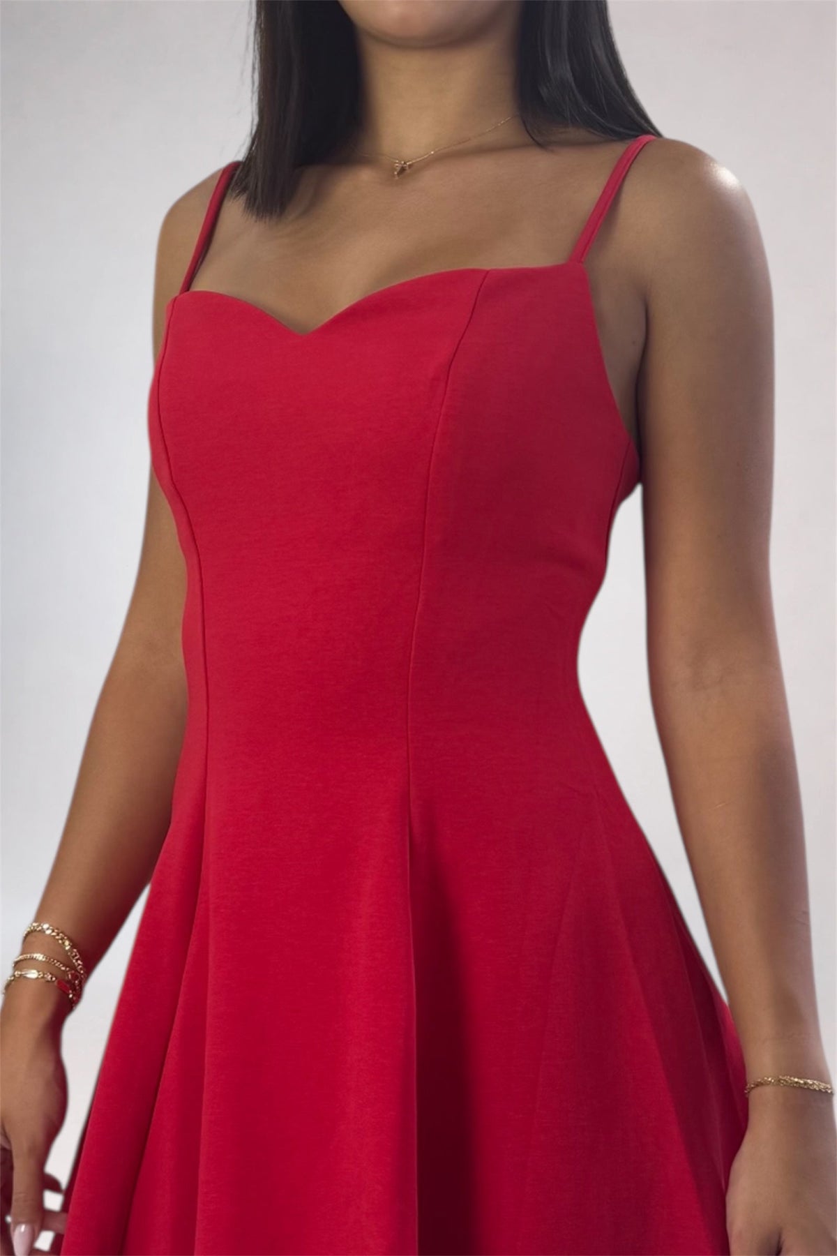 A woman wearing a bright red spaghetti-strap dress with a sweetheart neckline and a flared skirt, shown from the shoulders to the hips against a light gray studio background.