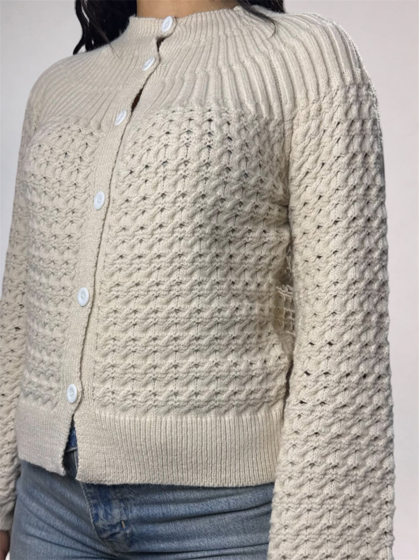 A close-up of a person wearing a cream-colored textured knit cardigan with white buttons, paired with blue jeans.