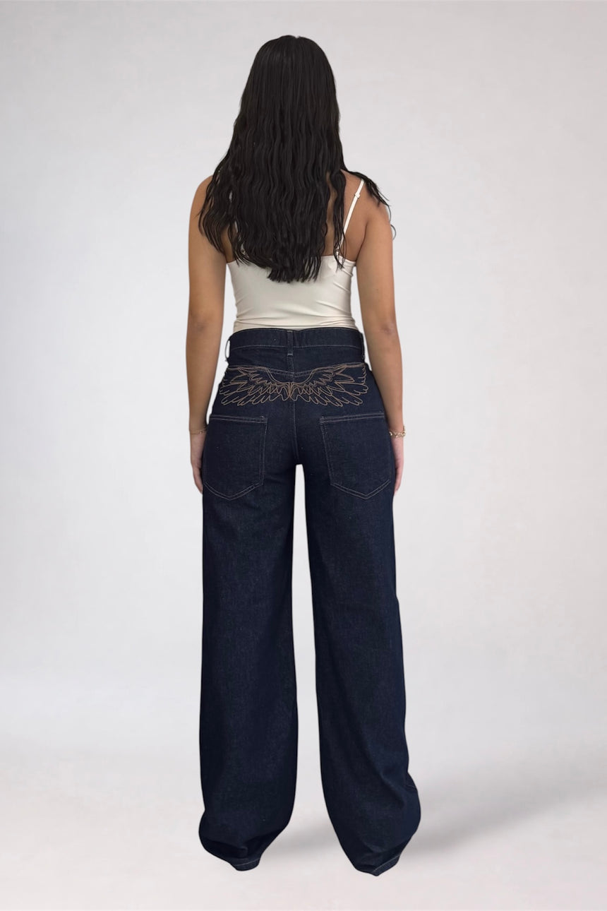 A woman viewed from behind wearing dark wide-leg denim jeans with a decorative embroidered pattern across the back waistband, paired with a white spaghetti-strap tank top, against a light gray studio background.