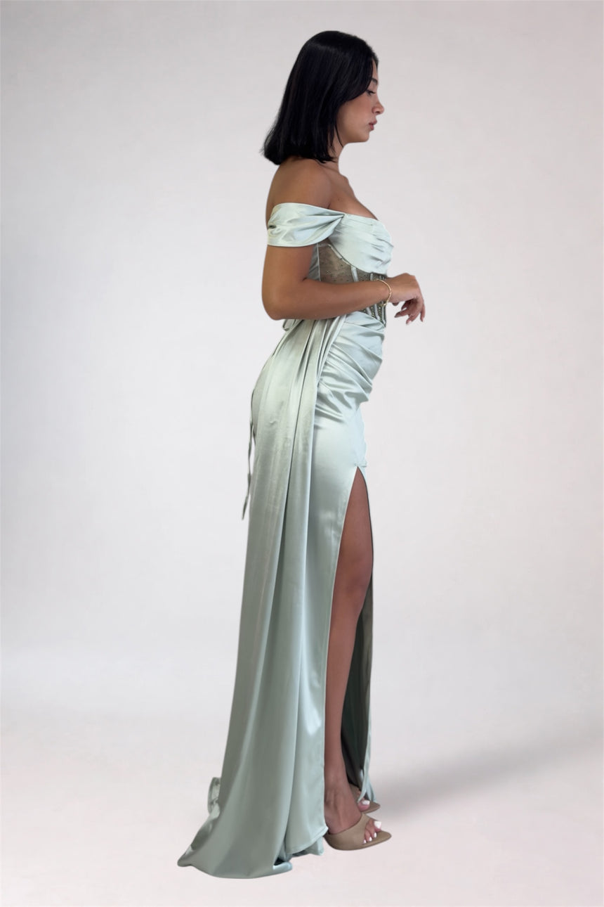 A woman modeling an off-the-shoulder floor-length gown in a pale mint green satin fabric, featuring a high thigh slit, against a plain light gray studio background.