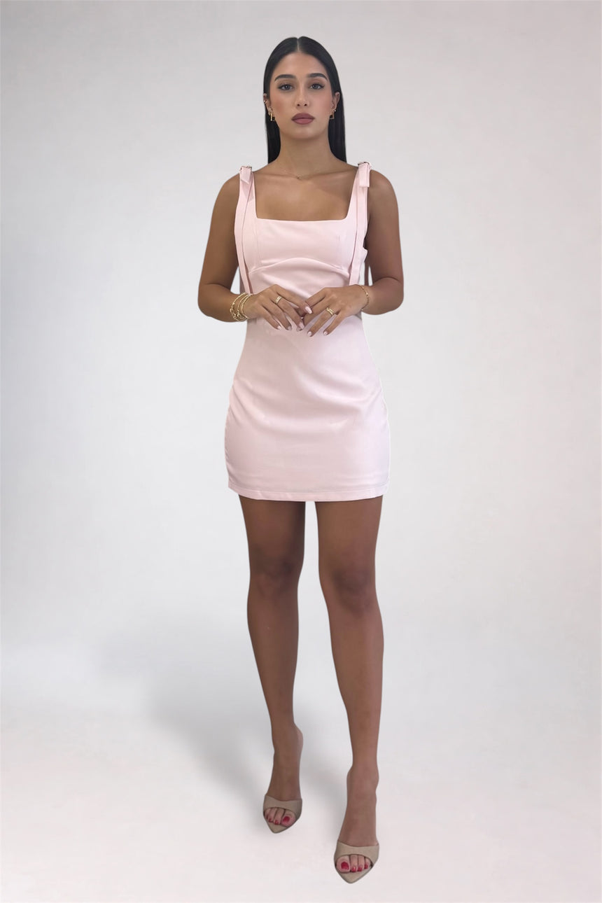 A woman modeling a light pink sleeveless mini dress with square straps and a gathered waist, paired with nude high heels, against a plain light gray studio background.