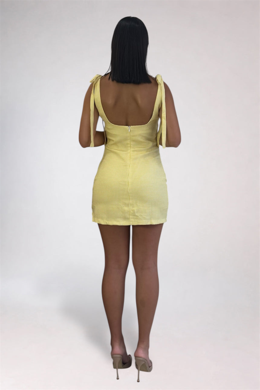 A woman viewed from behind wearing a pale yellow ribbed mini dress with tie-shoulder straps and a low open back, paired with nude high heels, against a light gray studio background.