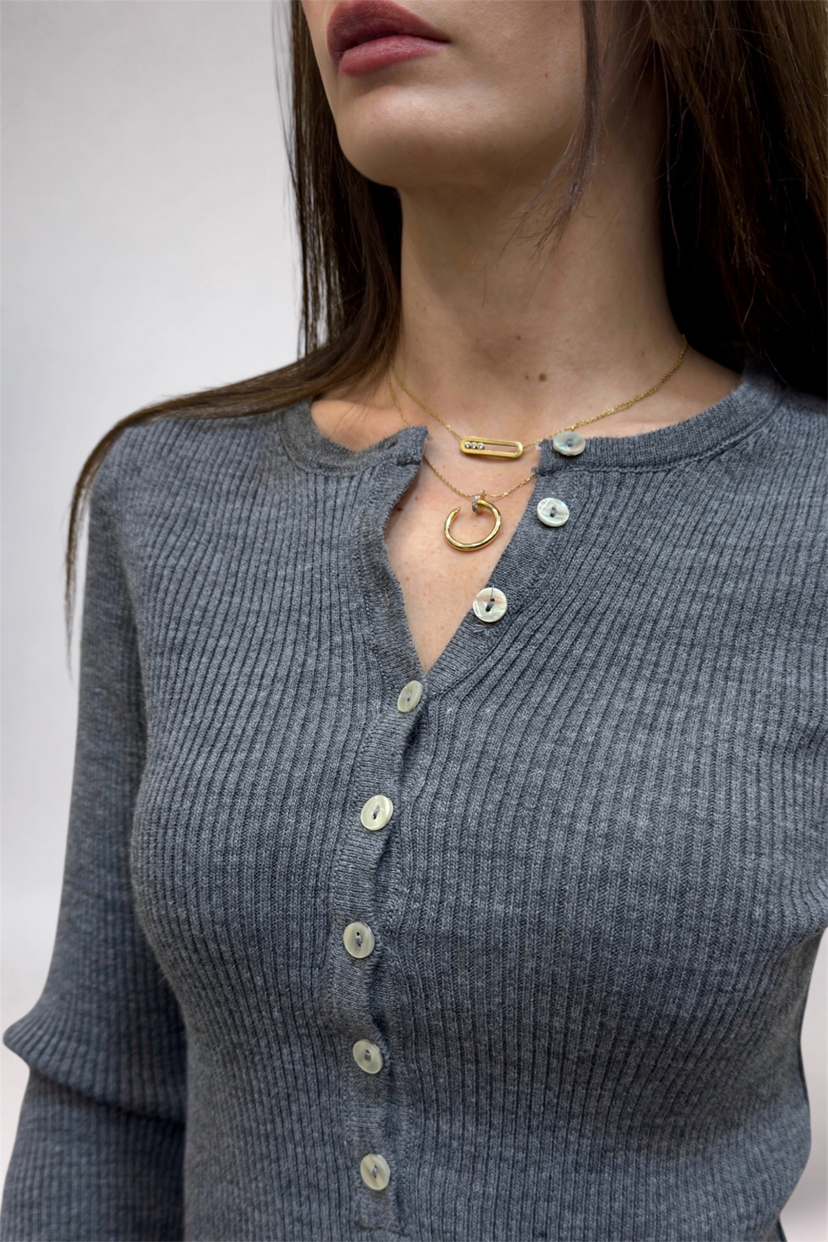 Woman wearing a gray ribbed sweater with buttons and gold necklaces against a neutral background