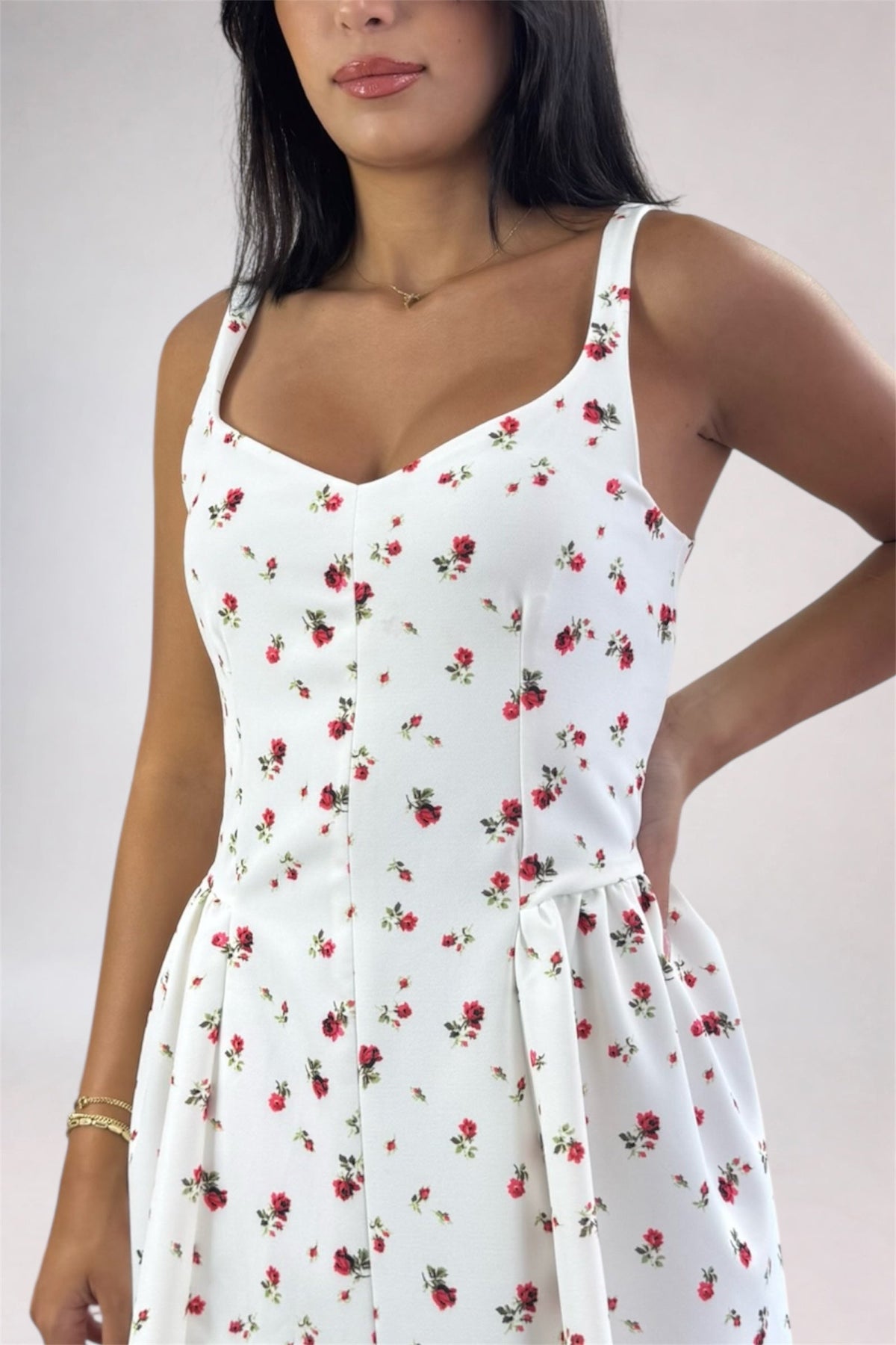 A woman wearing a white sleeveless dress with a red and green floral print, featuring a sweetheart neckline, thin shoulder straps, and a tie waist.